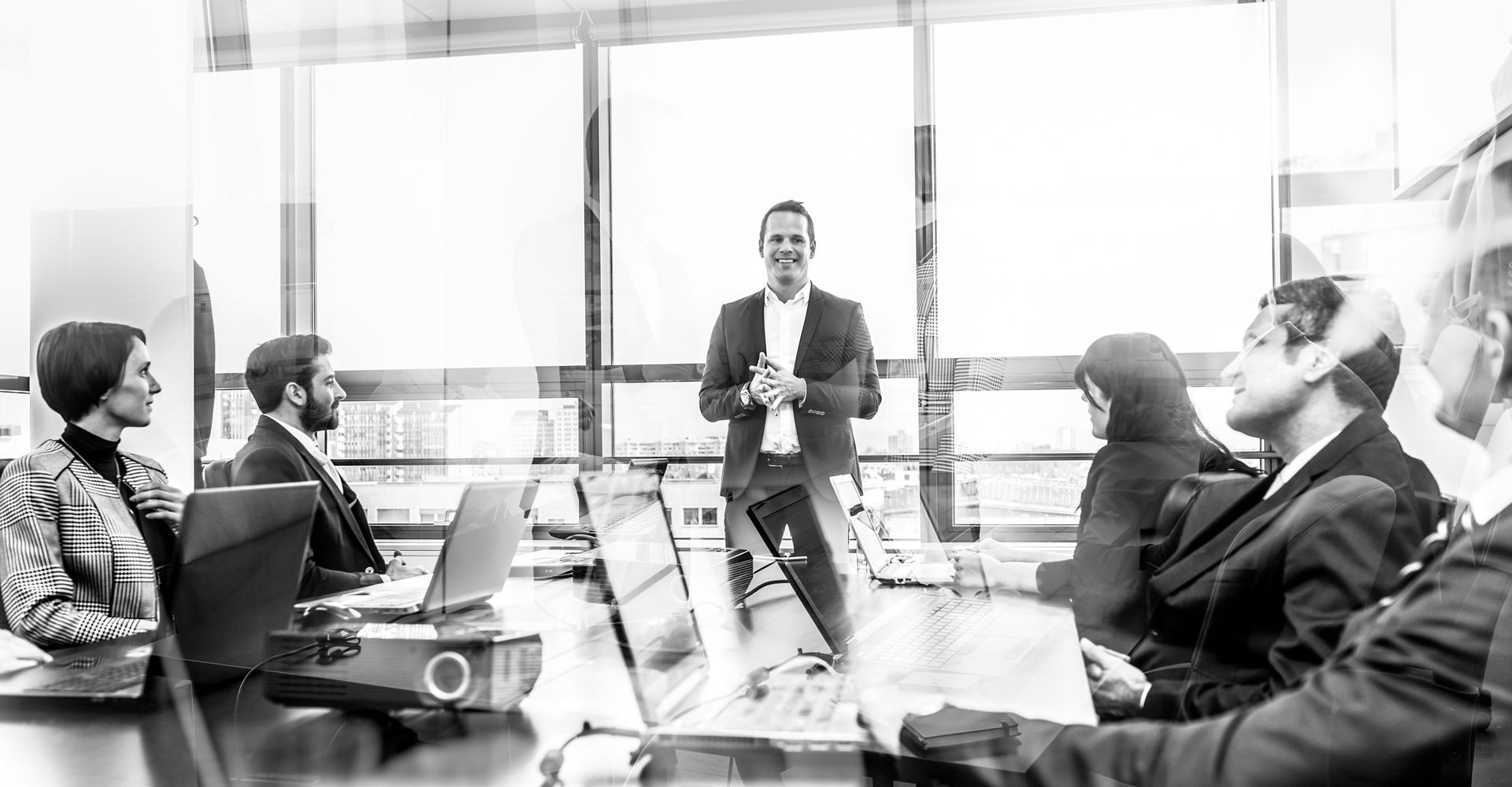 Successful team leader and business owner leading informal in-house business meeting. Businessman working on laptop in foreground. Business and entrepreneurship concept. Blue toned grayscale.